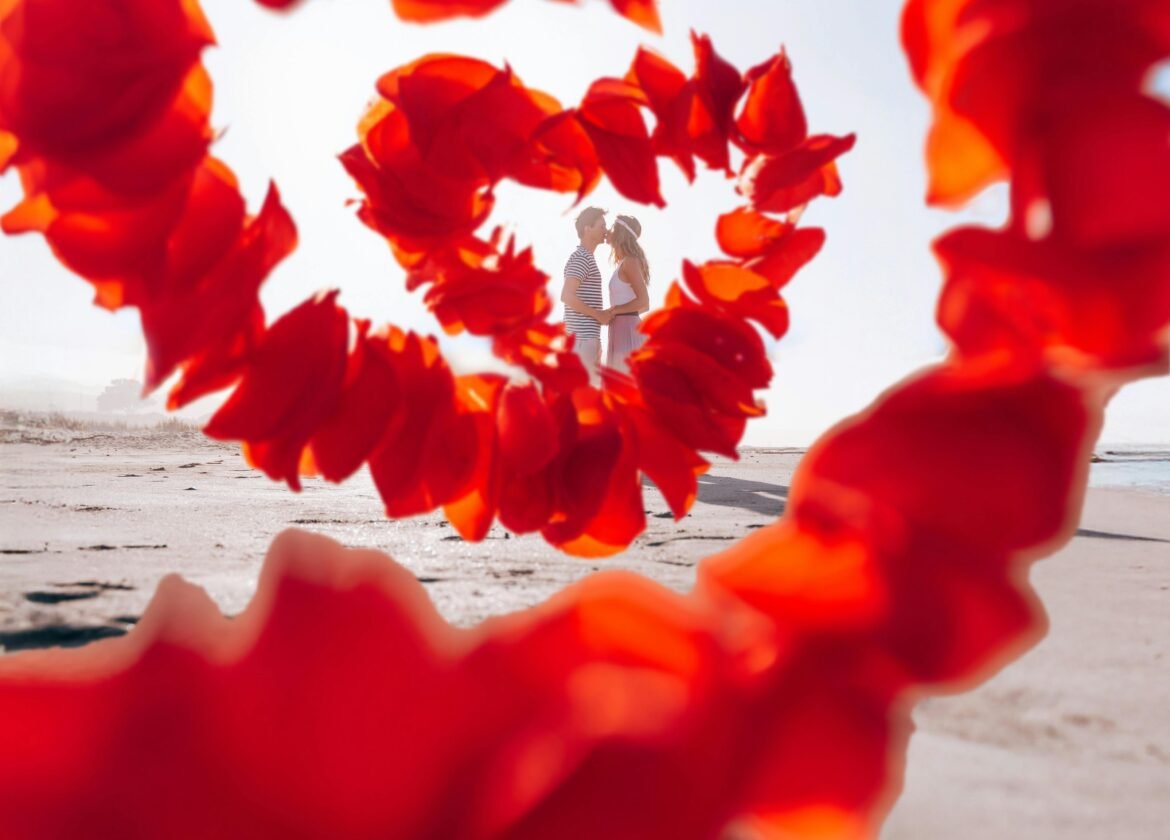 A Couple Embraces at the Beach, Framed by Vibrant Red Petals Forming A Heart Shape.