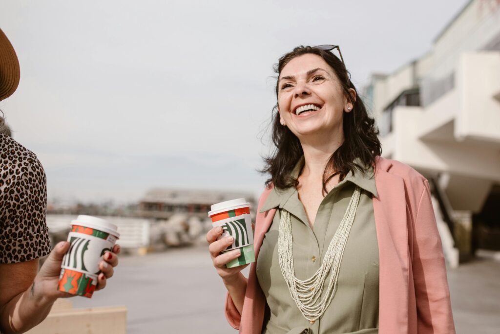 Smiling women enjoying conversation and coffee outdoors on a sunny day, promoting connection and lifestyle.
