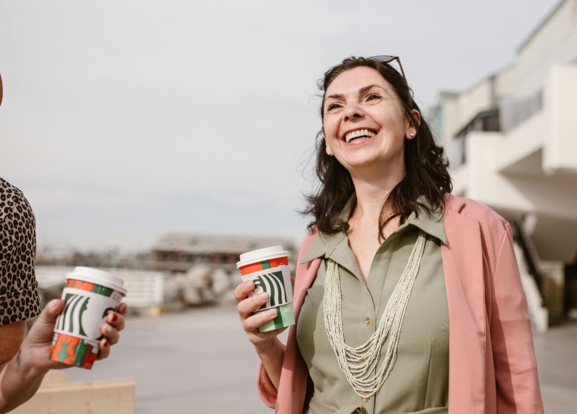 Smiling women enjoying conversation and coffee outdoors on a sunny day, promoting connection and lifestyle.