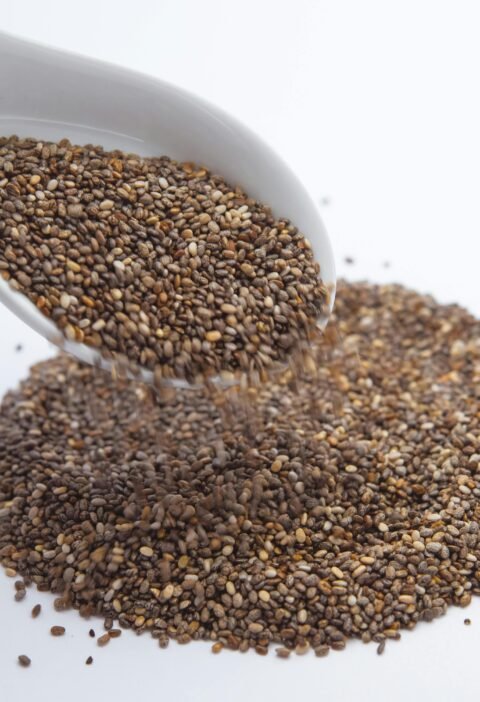 A close-up shot of chia seeds pouring from a white ceramic spoon onto a white surface.