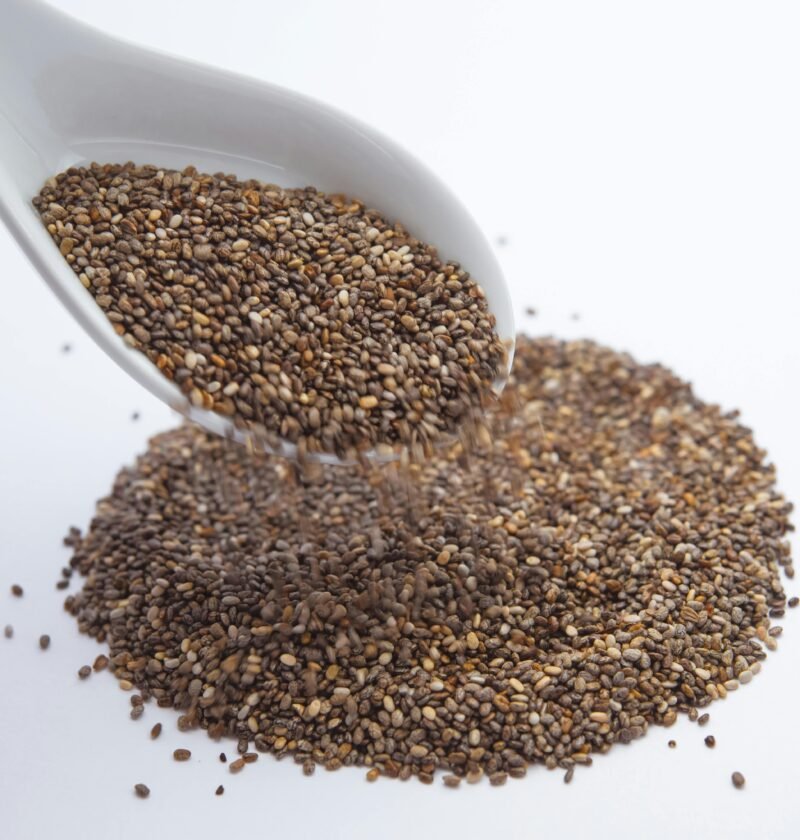 A close-up shot of chia seeds pouring from a white ceramic spoon onto a white surface.